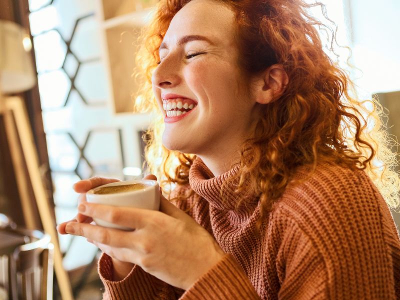 Mulher ruiva toma café e sorri. Imagem: Nastasic de Getty Images Signature - Canva.